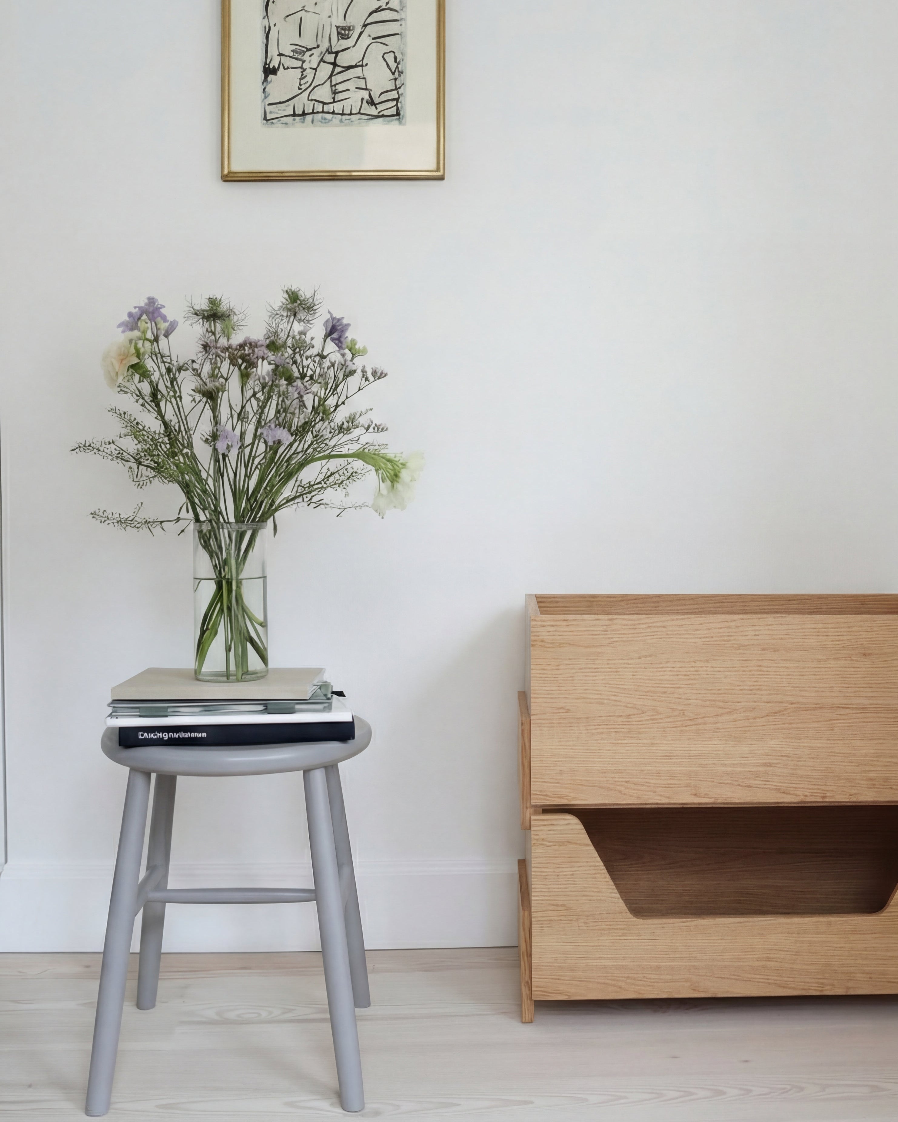 2 light natural oak wooden stackable storage  modules with a lower cutout , styled in a minimalist  scandinavian room next to a gray stool holding books and a vase of flowers.